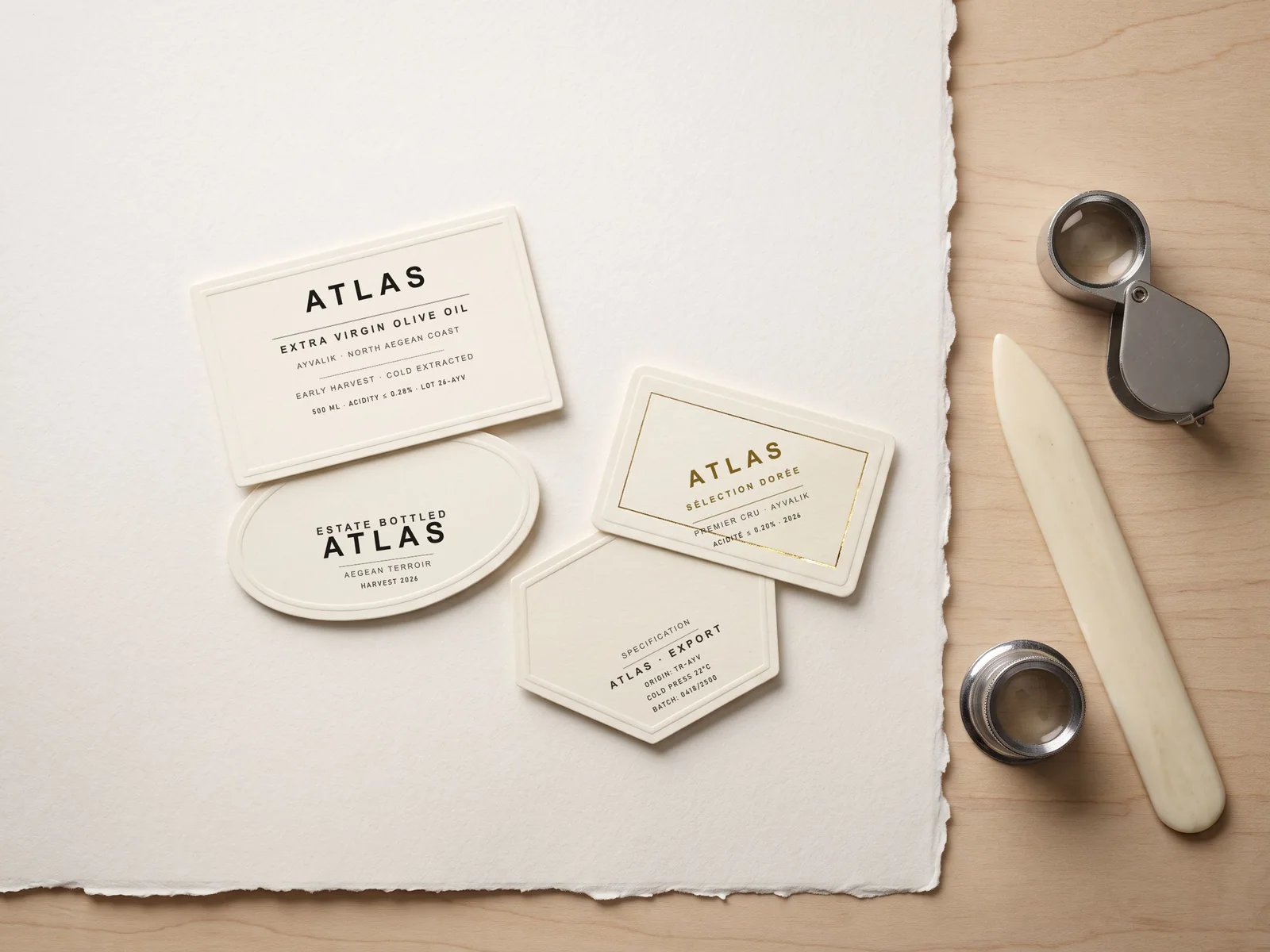 Overhead flat-lay photograph of four Atlas label die-cuts on heavy cotton paper with loupe and bone folder